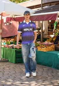 Dark grey and purple striped short-sleeve football jersey with white collar and logo, paired with distressed light blue jeans and white sneakers.