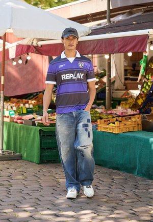 Dunkelgrau und lila gestreiftes Kurzarm-Fußballtrikot mit weißem Kragen und Logo, kombiniert mit ausgewaschenen hellblauen Jeans und weißen Sneakers.