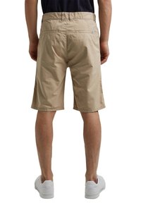 Beżowe bawełniane szorty o prostym kroju, z dwiema tylnymi kieszeniami i jednolitą powierzchnią, noszone z białymi sneakersami. Widoczne szwy na całej długości.