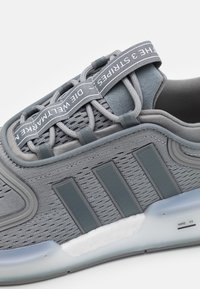 Grå Adidas-sneaker med ovandel i mesh, tre svarta ränder på sidan, elastiska snören och en genomskinlig vit sula märkt NMD V3.