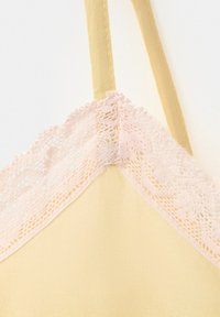 Vêtement en tissu jaune avec bordure en dentelle rose pâle et fine bretelle, détail en gros plan sur l'encolure et l'attache de la bretelle.