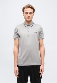 Νέος άνδρας με ανοιχτό καστανά μαλλιά που φοράει ανοιχτό γκρι πόλο μπλουζάκι με το λογότυπο «BOSS» στο στήθος κα ι μαύρο παντελόνι, στέκεται μπροστά σε απλό φόντο.