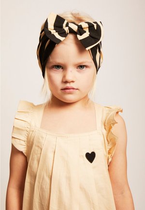 Black and beige striped fabric headband with a bow design. Light beige dress with ruffled sleeves and a small black heart patch.