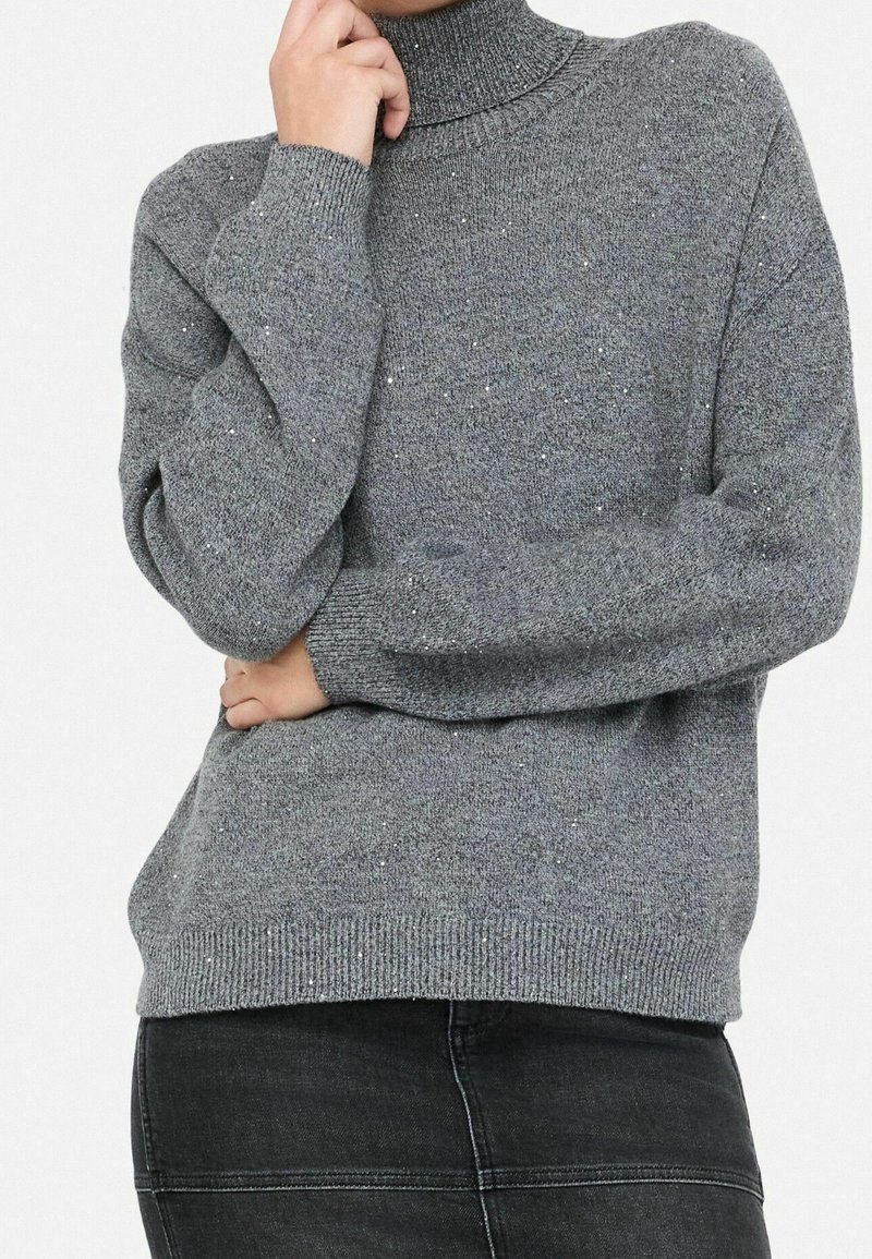 Personne portant un pull à col roulé gris moucheté et un pantalon noir, une main touchant son visage et l'autre croisée.