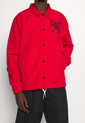 Homme portant une veste rouge à boutons avec du texte et des logos noirs "CHI" sur la poitrine et un pantalon noir avec des cordons blancs.