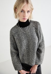 Maglione a coste con motivo a chevron bianco e nero, con colletto alto nero a costine e spalle cadenti, caratterizzato da una finitura testurizzata.