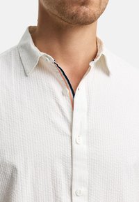 Camisa blanca texturizada con cuello abotonado, que presenta un ribete interior en contraste en azul marino y naranja, y botones blancos.