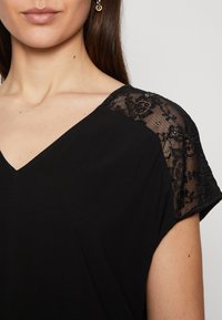 Svart topp med V-hals og korte ermer, pyntet med blonder i blomstermønster på skuldrene, glatt tekstur.