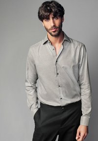 Cămașă cu carouri alb-negru, cu guler, mâneci lungi și un singur buzunar pe piept, purtată cu pantaloni negri. Textură de țesătură netedă.