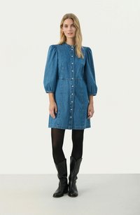 Denim dress in blue with puffed sleeves and buttons down the front, featuring a fitted waistline and a flared hem. Worn with black boots.
