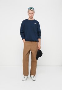 Navy blue sweatshirt with logo, tan wide-leg pants, white sneakers with multicolored accents, holding a black cap. Smooth texture, casual design.
