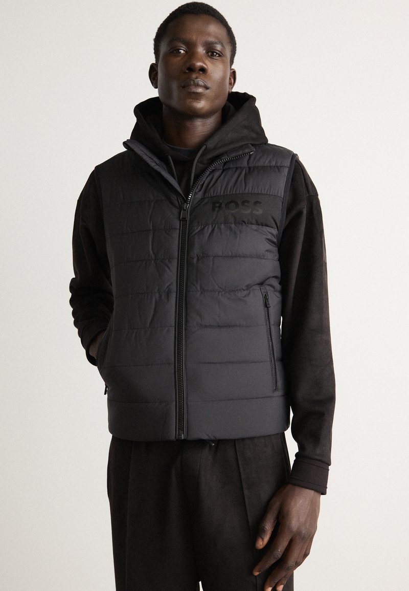 Black padded vest with horizontal stitching, front zipper, and subtle branding. Worn over a black hoodie, featuring high collar and kangaroo pockets.