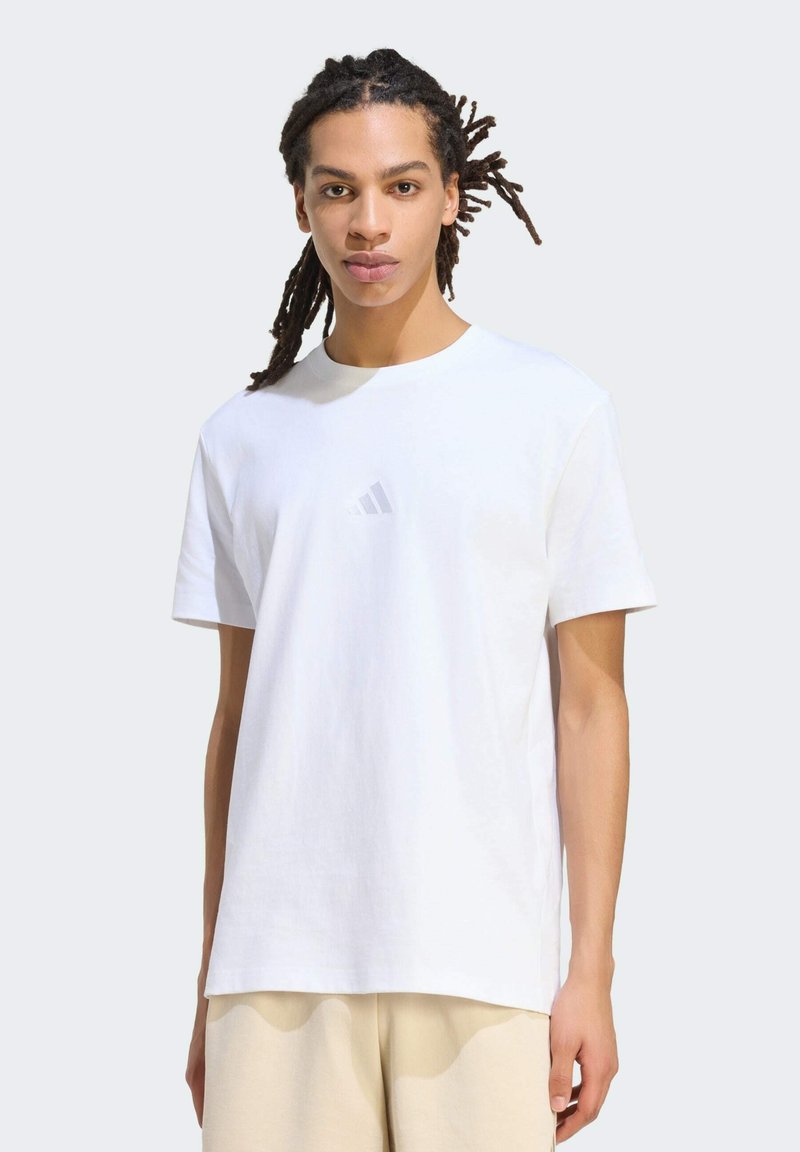 Jeune homme aux dreadlocks portant un t-shirt blanc uni Adidas et un pantalon beige, debout devant un fond gris clair.