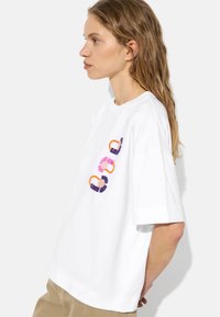 Biała bawełniana koszulka z krótkim rękawem, zdobiona kolorowymi haftowanymi wzorami w odcieniach różu, fioletu i pomarańczowego z przodu.