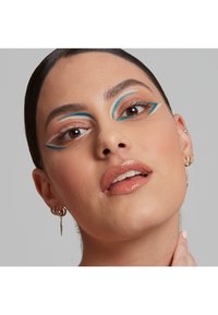 Närbild av ett ansikte med dramatisk sminkning som har blå och vita geometriska mönster runt ögonen. Skinande läppar och guldörhängen syns.