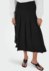 Žena koja nosi crnu plisiranu midi suknju, bijelu bluzu dugih rukava, crne sandale s potpeticom i drži malu crnu torbicu.