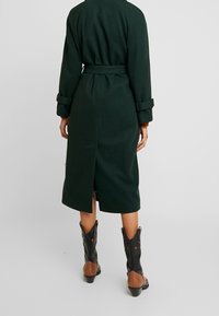 Pitkä vihreä takki, jossa on vyötäröllä vyö, käännetyt hihat ja takana halkio; yhdistetty mustiin ja ruskeisiin cowboy-saappaisiin, joissa on tähtikoristeita.