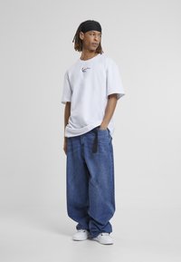 T-shirt blanc à manches courtes avec un graphique noir, associé à un jean en denim bleu ample avec une étiquette noire. Des baskets blanches complètent la tenue.