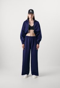 Giacca cropped blu navy con polsini elasticizzati, abbinata a pantaloni larghi coordinati. Reggiseno sportivo nero visibile sotto. Sneakers bianche indossate.