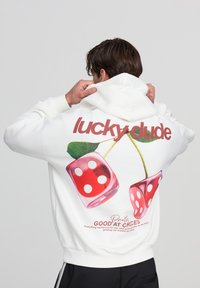 Felpa bianca con una grafica di dadi rossi e foglie verdi. Il testo "fortuna amico" e "Abilità nelle scelte" stampato in rosso e nero.