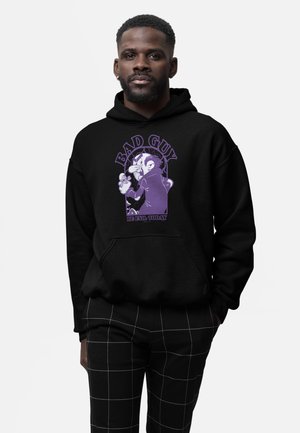 Mężczyzna ubrany w czarną bluzę z kapturem z fioletową postacią z rysunku i napisem „Bad Guy Be Evil Today”, w połączeniu z czarnymi spodniami z białymi liniami kratki.