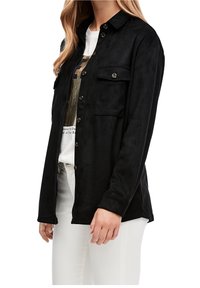 Donna con una camicia giacca nera con bottoni e tasche a patta sopra una t-shirt bianca con grafica e pantaloni bianchi, in piedi su uno sfondo bianco.