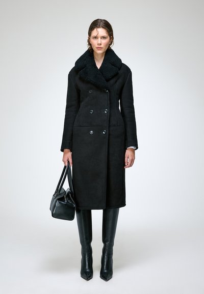 Femme portant un long manteau noir croisé avec un grand col, des bottes noires montantes, tenant un sac à main noir, debout devant un fond uni.