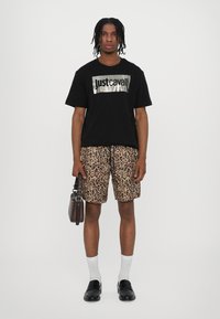 Zwarte t-shirt met korte mouwen en metallic logo, gecombineerd met leopardprint shorts. Heeft een bruine tas; sportieve witte sokken en zwarte schoenen.