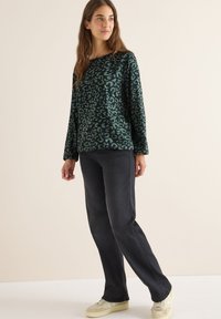 Maglietta a maniche lunghe di colore verde scuro con motivo maculato, abbinata a pantaloni neri e sneaker crema, con un fit rilassato.