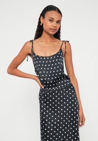 Zwarte satijnen jurk met witte polka dots, voorzien van spaghettibandjes en een losse pasvorm. De stof heeft een gladde, glanzende textuur.