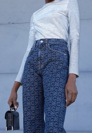 Personne portant un jean bleu taille haute avec un imprimé intégral du logo "MCM" et un haut à manches longues argenté, tenant un petit sac à main noir.