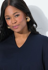 Tummansininen V-vetsyinen paita, jossa on sileä pinta; mallilla on yllään isot kultaset korvakorut; pehmeä valaistus korostaa kasvojen piirteitä.