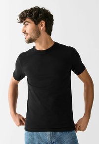 Vīrietis ar cirtainiem matiem, valkājot melnu īsroku T-kreklu un zilas džinsas, skatās uz sāniem un pielāgo krekla apakšu.