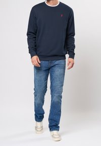 Sweat-shirt bleu marine à manches longues et poignets côtelés, arborant un petit logo rouge sur le côté gauche de la poitrine, associé à un jean bleu clair et des baskets.