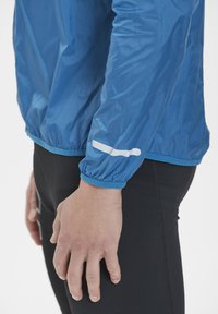 Ljusblå lättviktsjacka med elastiska ärmslut och ett kontrasterande vitt streck på ärmen, tillsammans med svarta tajta byxor.