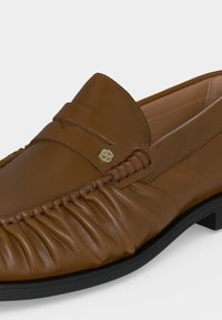 Mocassin en cuir marron avec côtés froncés, semelle noire et un petit emblème décoratif doré sur la partie supérieure.