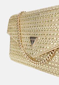 Auksinė rankinė su tekstūruota paviršiumi, geometriniais raštais ir trikampiu logotipu. Ji turi auksinę grandinėlę ir užsegimą su sklende.