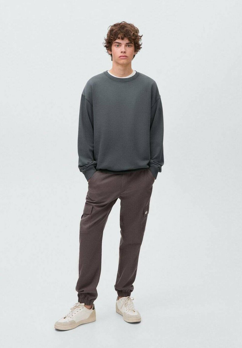 Felpa grigia con maniche lunghe, abbinata a pantaloni cargo marroni con tasche. Scarpe bianche con accenti beige.