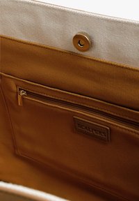 Interieur van een bruine stoffen tas met een gladde textuur, voorzien van een ritsvak en een gestikt label met het merk "SCALPERS." Gouden drukknoopsluiting.