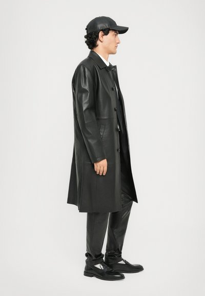 Man wearing a black leather long coat, black leather pants, black cap, and black loafers, standing sideways against a plain white background.