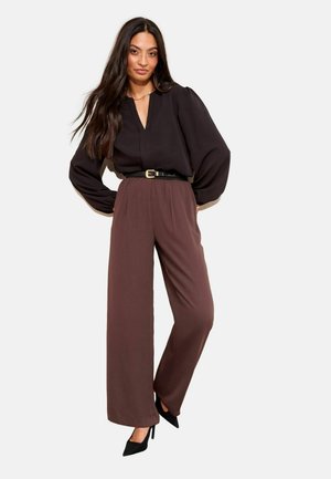 Frau mit langen dunklen Haaren, die eine schwarze Bluse mit langen Ärmeln, braune weite Hosen, einen schwarzen Gürtel und schwarze High Heels trägt, und mit auf die Hüften gestützten Händen steht.