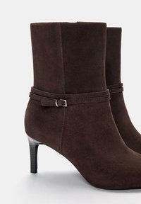 Stivali ankle in suede marrone con design aderente, punta affusolata e tacco alto sottile. Presentano una cintura decorativa con una fibbia argentata.
