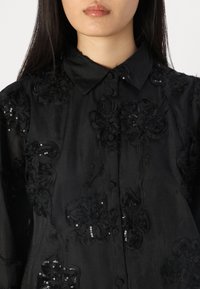 Camisa preta com bordados florais em lantejoulas, design abotoado e textura suave. A gola e os padrões detalhados são proeminentes.