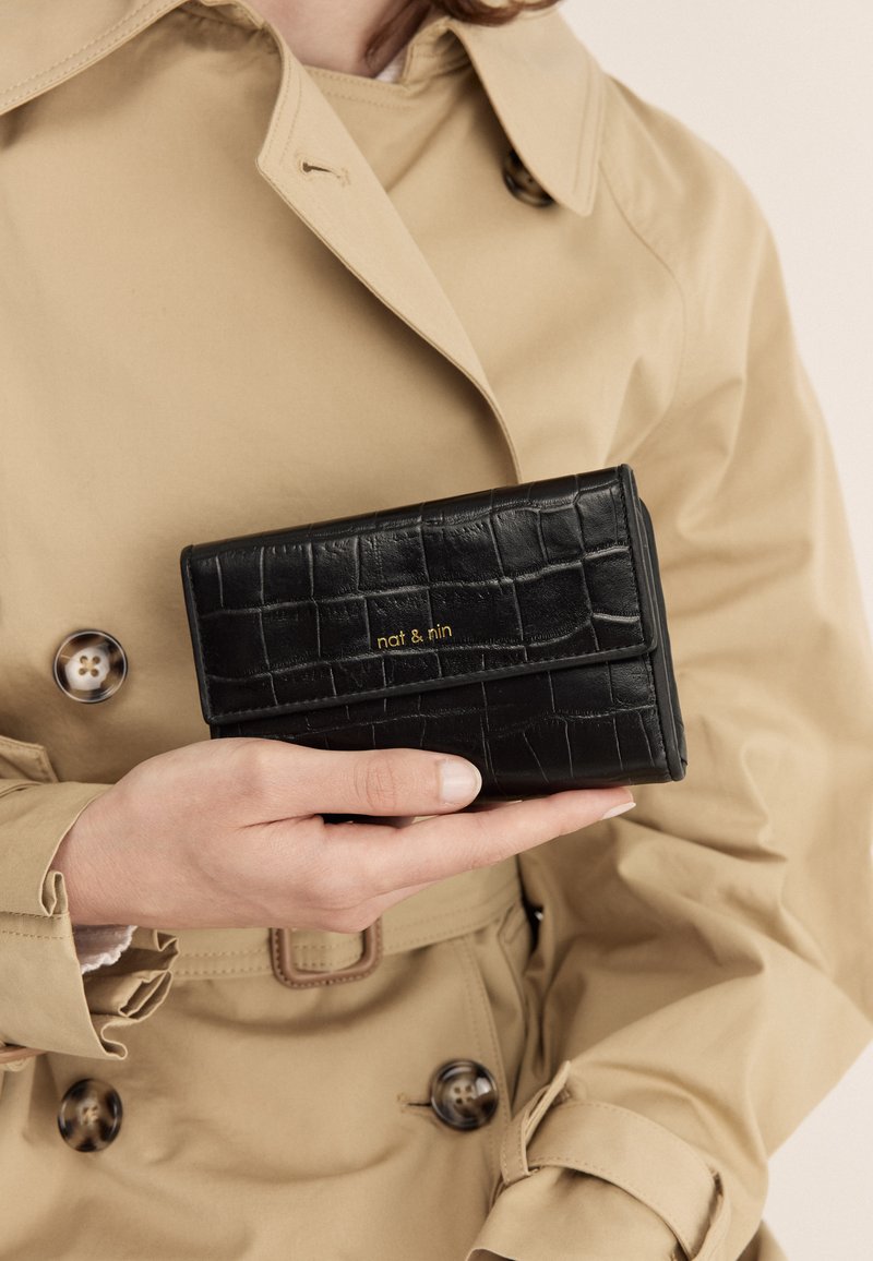 Portafoglio in pelle nera con stampa coccodrillo, con chiusura a patta e dettagli in oro. Tenuto in mano su uno sfondo di trench beige.