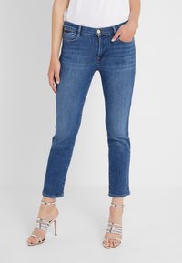 Jeans de mezclilla ajustados en azul oscuro con cintura de altura media y un sutil desgaste; combinados con sandalias de tacón con tiras plateadas.