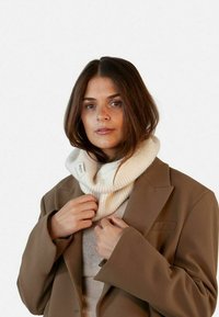 Sciarpa a coste di colore crema indossata attorno al collo, abbinata a un blazer oversize beige. La sciarpa presenta un'etichetta su un lato.