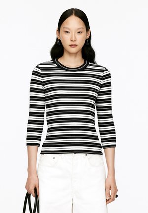 Woman with long black hair wearing black and white striped three-quarter sleeve top and white pants, holding a black handbag.