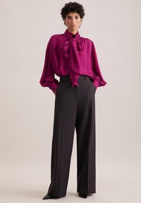 Blusa di seta fucsia con un grande fiocco al colletto, abbinata a pantaloni neri a gamba larga con vita alta, che mostrano trame lisce e sartorialità.