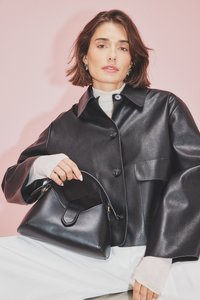 Čierna kožená bunda s veľkými gombíkmi a bočnými vreckami, skombinovaná s malou čiernou kabelkou so zakriveným dizajnom a zlatými kovovými doplnkami.