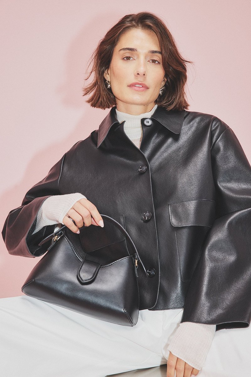 Čierna kožená bunda s veľkými gombíkmi a bočnými vreckami, skombinovaná s malou čiernou kabelkou so zakriveným dizajnom a zlatými kovovými doplnkami.